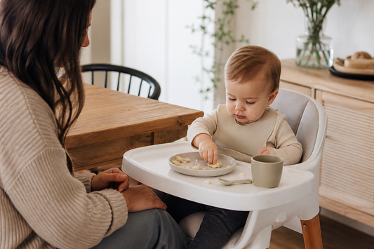 Caregiver supervising a baby during a simple self-feeding meal with minimal tableware